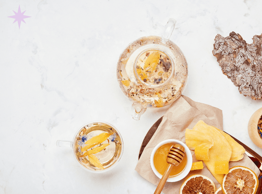 tea flat lay A flat lay of a glass teapot and teacup filled with floral herbal tea on a white marble surface. Next to them are dried orange slices, a bowl of honey with a dipper, and pieces of dried mango on parchment paper, creating a cozy, natural tea-time scene.