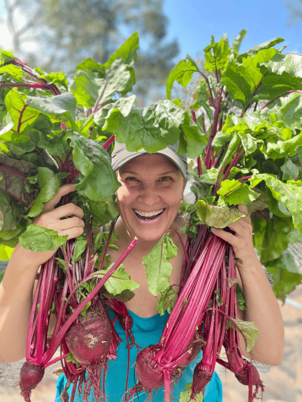 candace beets A smiling woman in a teal tank top and gray cap holds up large, freshly harvested bunches of beets with vibrant green leaves and deep red roots. She stands outdoors in the sunshine, radiating joy and connection to the garden.
