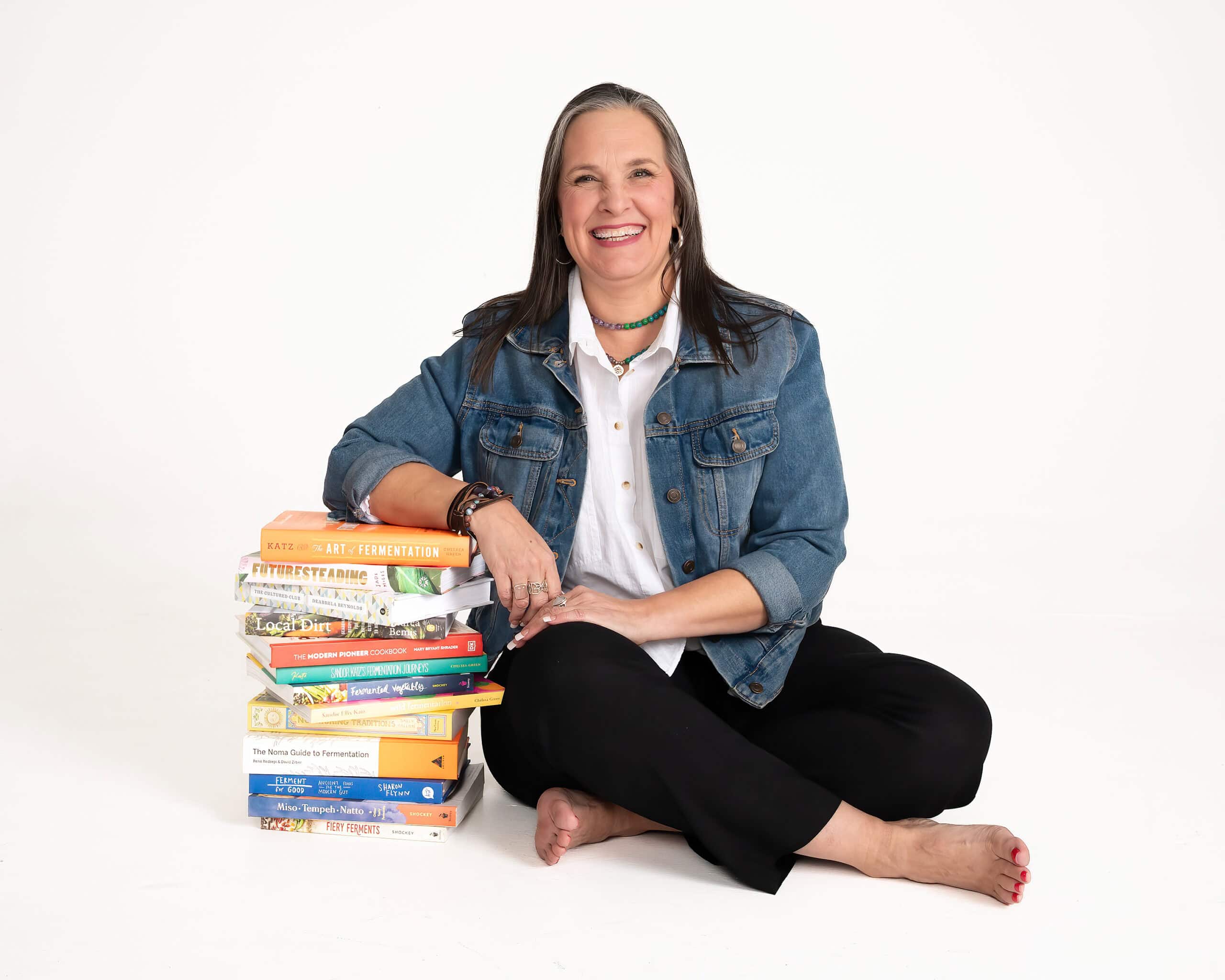 A smiling woman with long hair sits barefoot on the floor against a white backdrop. She wears a denim jacket over a white shirt and black pants, resting one arm casually on a tall stack of colorful books about fermentation, gardening, and sustainable living.
