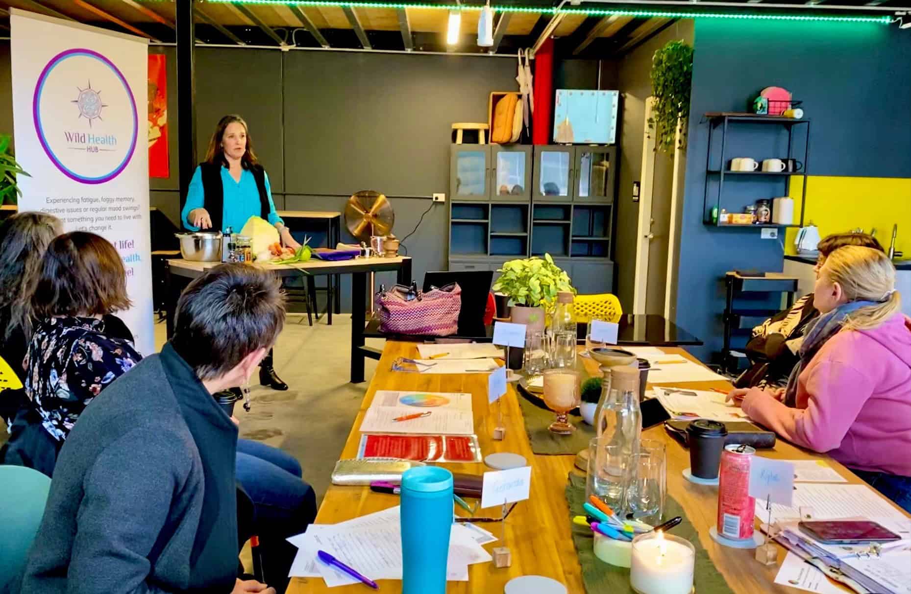 Presentation 2 A woman stands at the front of a modern workshop space, leading a Wild Health Hub presentation with fresh produce and fermentation tools on the table beside her. Seated around a long wooden table, participants listen attentively, with papers, colorful pens, candles, and drinks spread out in front of them, creating a cozy and engaged learning atmosphere.