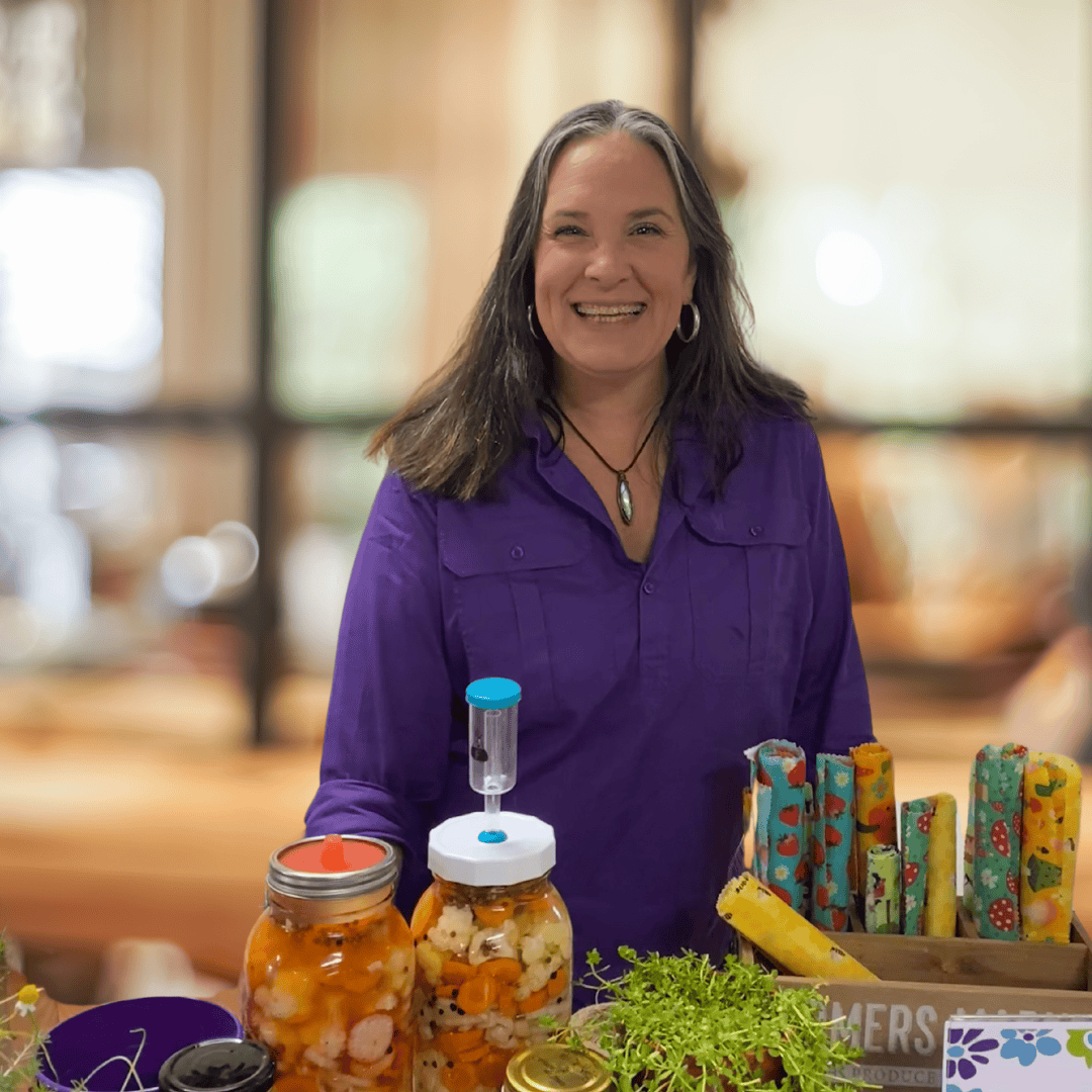 Untitled design A smiling woman in a purple shirt stands behind a colorful display table featuring jars of fermented vegetables, fresh sprouts, and a crate of rolled beeswax wraps with playful prints. The setting appears to be an indoor market or workshop space with a warm, softly lit background.