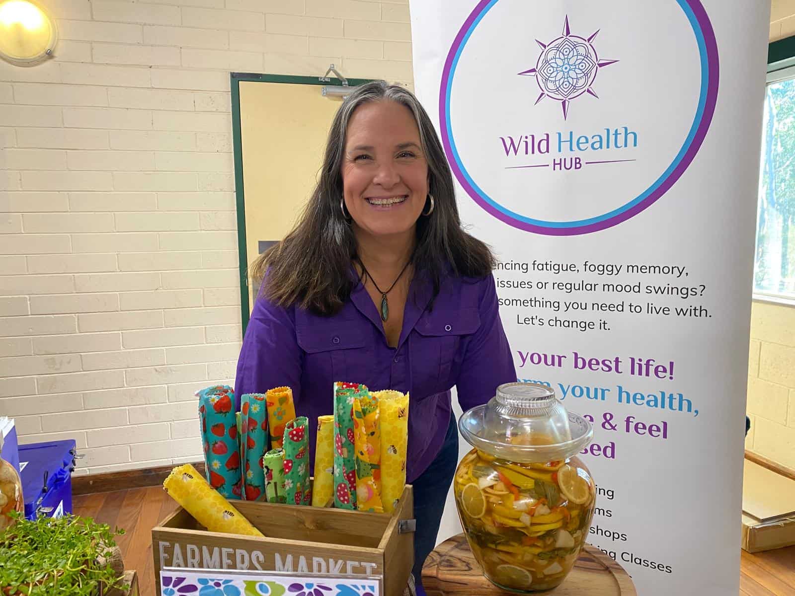 candace markets A woman in a purple shirt smiles while standing behind a market-style display at a Wild Health Hub booth. In front of her are colorful beeswax wraps in a wooden crate labeled “Farmers Market” and a large glass jar filled with vibrant citrus peels and vegetables in liquid, likely a ferment or infusion; a branded banner promoting health and wellness stands in the background.
